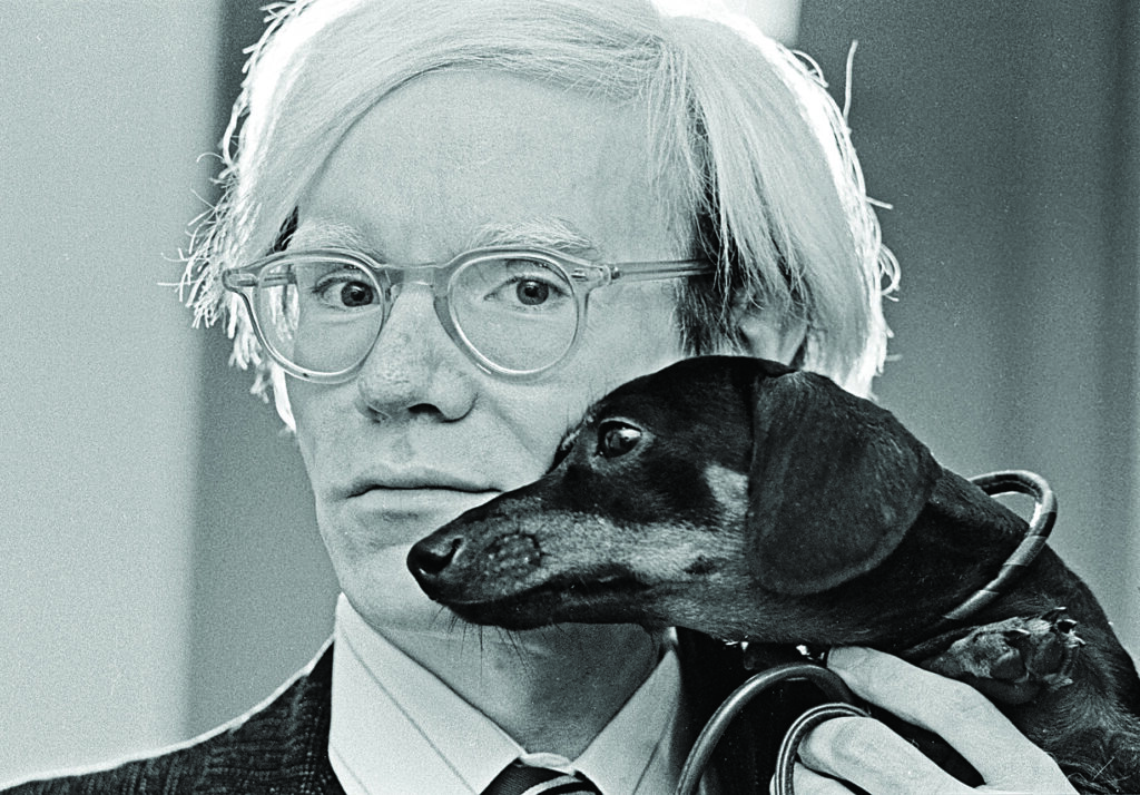 a man in glasses and a suit holds a small dog affectionately so the dog's profile is next to the man's face