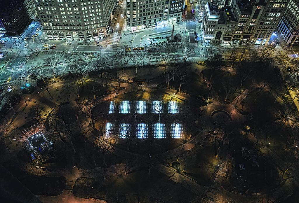 an aerial, birds-eye, nighttime view of an urban park surrounded by city streets and tall buildings, with illuminated rectangular light installations arranged on the lawn among leafless trees