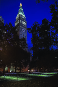 lluminated clock tower rising above a city park at night, framed by dark trees, with small lights scattered across the grass as part of an art installation glow beneath a deep blue sky