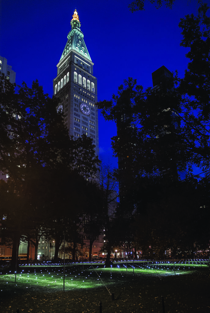 lluminated clock tower rising above a city park at night, framed by dark trees, with small lights scattered across the grass as part of an art installation glow beneath a deep blue sky