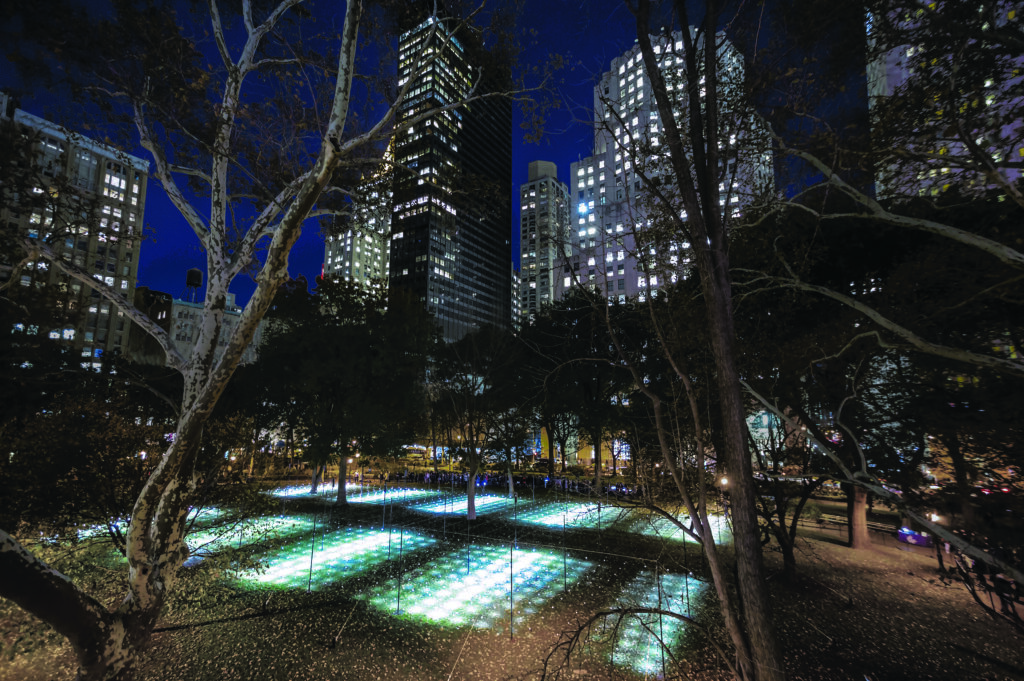 Nighttime view of an urban park surrounded by city streets and tall buildings, with illuminated rectangular light installations arranged on the lawn among leafless trees
