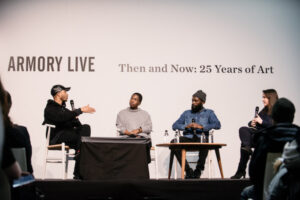 four people on a stage with microphones engage in conversation during a live podcast