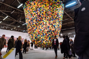 people walk through a large gallery space with a large multicolored sculpture suspended in the air from an industrial ceiling