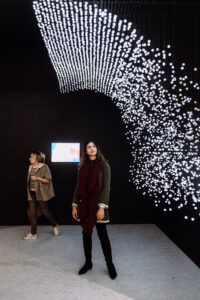 A woman looks up at a light sculpture in dark gallery