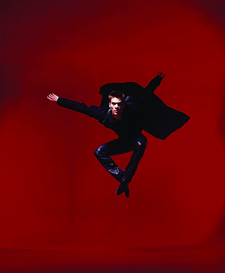 A dancer in an all-black costume with a cape leaps into the air against a dramatic red background