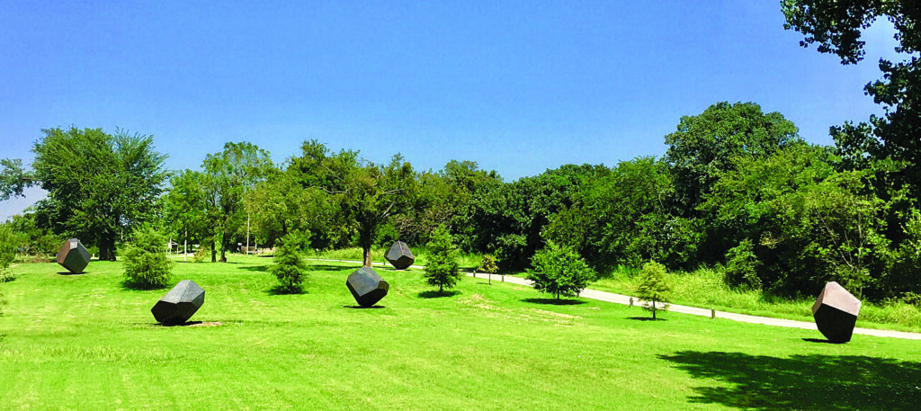 A grassy lake park dotted with large dark geometric sculptures