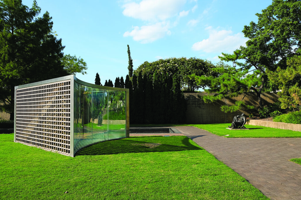 A landscaped outdoor area featuring a modern glass-and-metal structure beside a seated human‑figure sculpture
