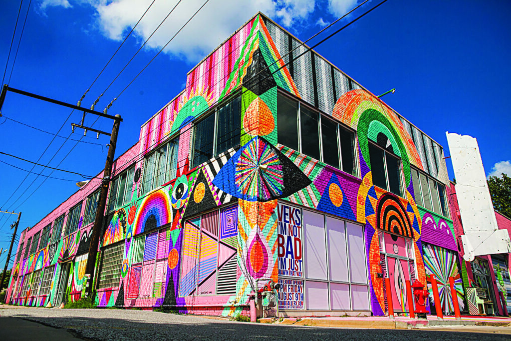 Corner view of a two-story industrial building covered in a colorful geometric mural with bright circles, arches, stripes, and abstract patterns against a blue sky