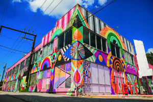 Corner view of a two-story industrial building covered in a colorful geometric mural with bright circles, arches, stripes, and abstract patterns against a blue sky