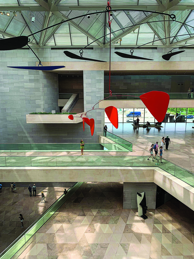 A spacious modern museum atrium where visitors move along multiple levels beneath a large red‑and‑black mobile