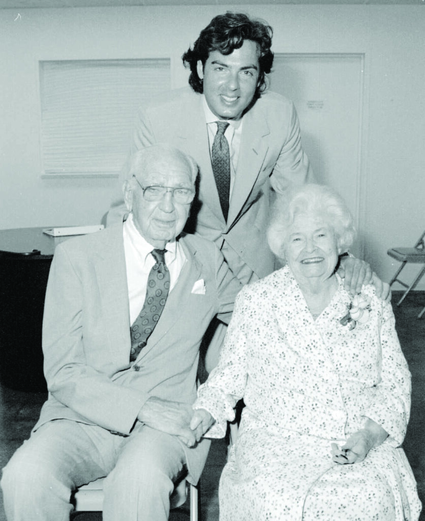 an elderly couple poses with their adult grandson at a party in honor of the grandmother