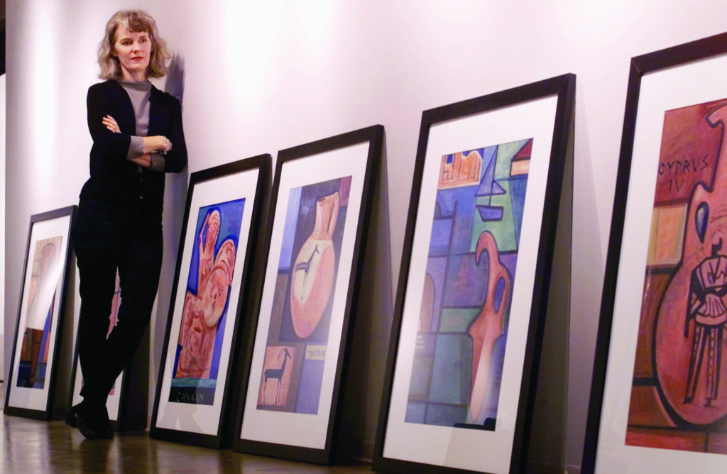 Artist Susan Morrison looks at some samples of her artwork as she and the artworks lean against a blank wall