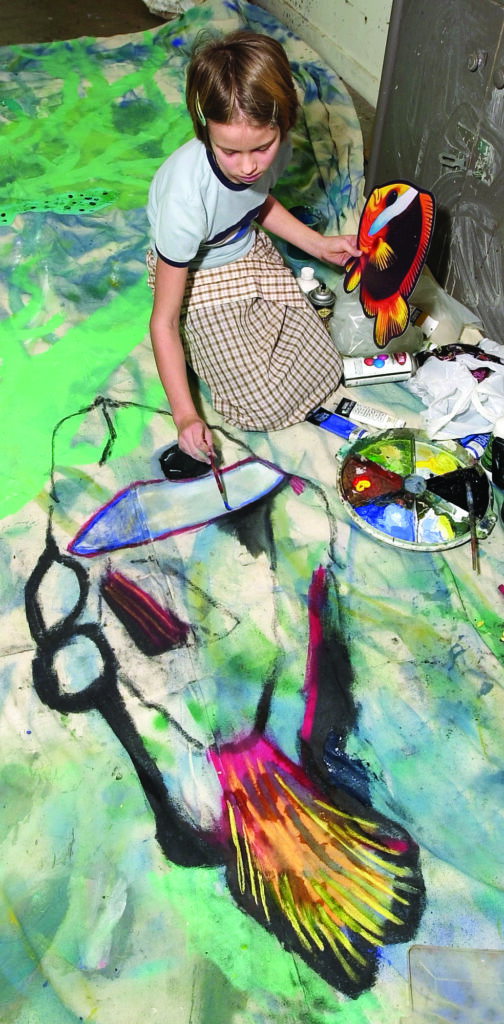 a girl sits on the ground and paints a fish on a large scale canvas at spring break art camp themed "Under the Sea"