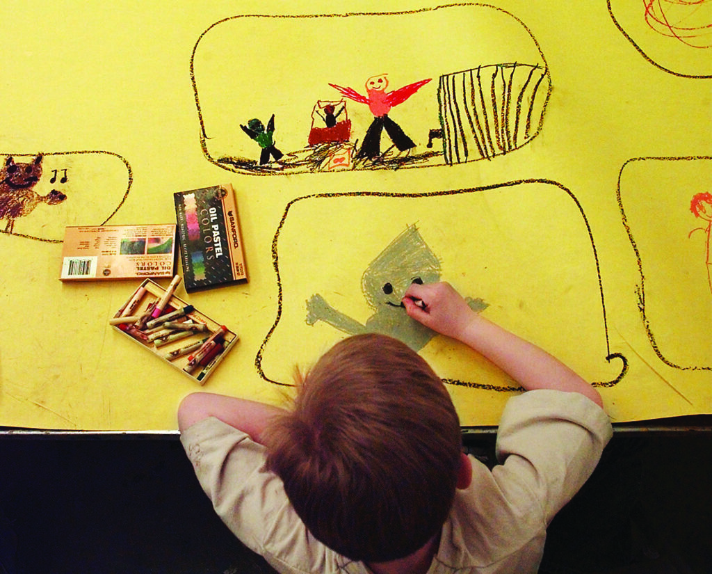 a child draws and colors in a defined space on a large roll of yellow paper as part of a collaboration creating scenes from "The Wizard of Oz" on a yellow brick road at art camp