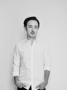 black and white portrait of composer Adam Crystal, who stands against a blank white wall in a white shirt with his hands in pockets as he looks down and to the left