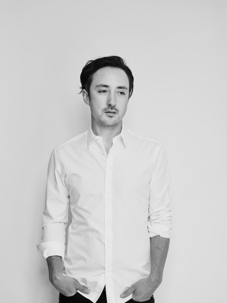 black and white portrait of composer Adam Crystal, who stands against a blank white wall in a white shirt with his hands in pockets as he looks down and to the left