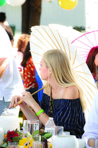 a woman shaded by a white aesthetic umbrella looks toward a speaker at a garden party