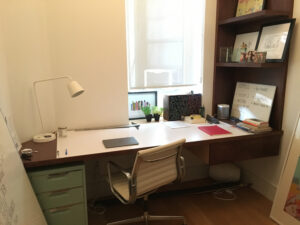 A desk scattered with notebooks is flanked by a bookshelf against a small, windowed wall in a New York City apartment