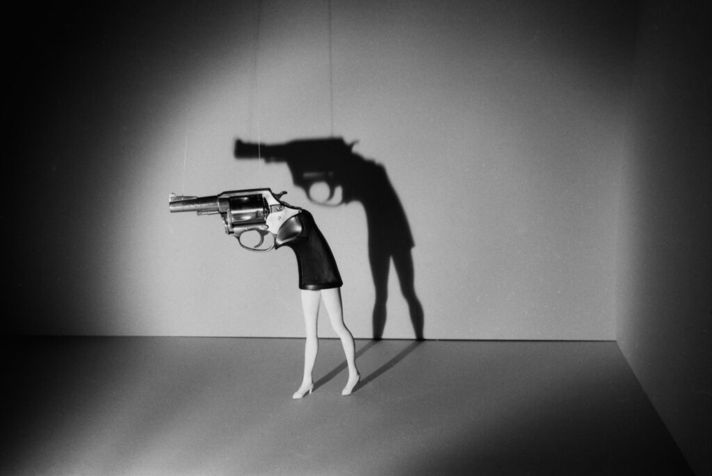 the legs and torso of a woman transform in the barrel of a gun with a center-stage spotlight reflecting the shadow of the half-woman, half-gun in the background