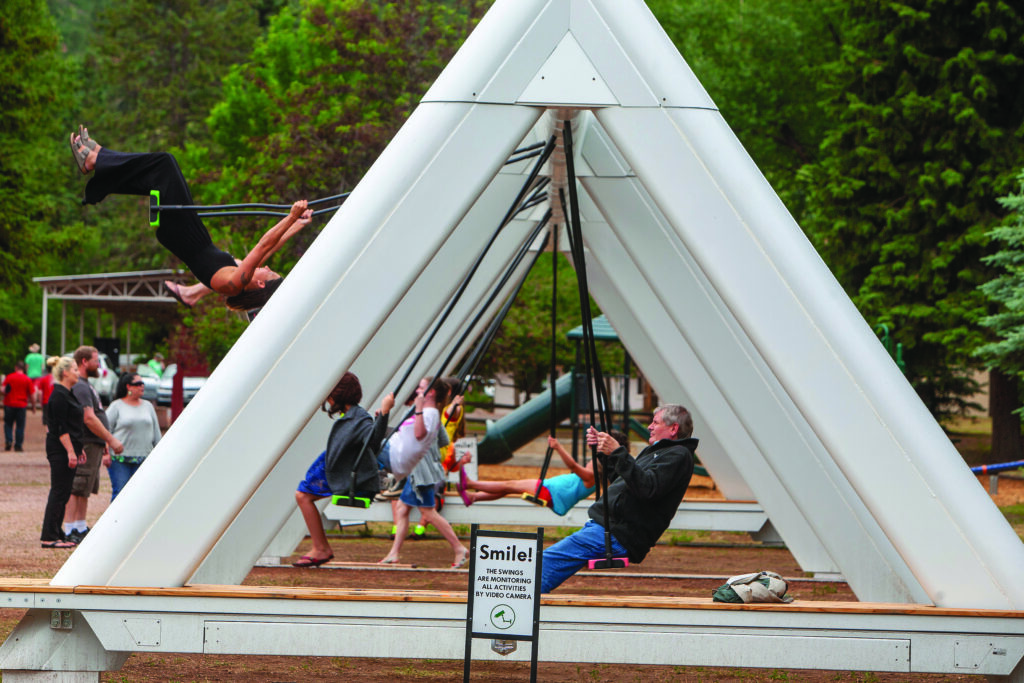 People using a large outdoor swing installation supported by triangular metal frames, with one person swinging upside down while others swing or watch nearby.