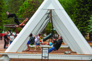 People using a large outdoor swing installation supported by triangular metal frames, with one person swinging upside down while others swing or watch nearby.