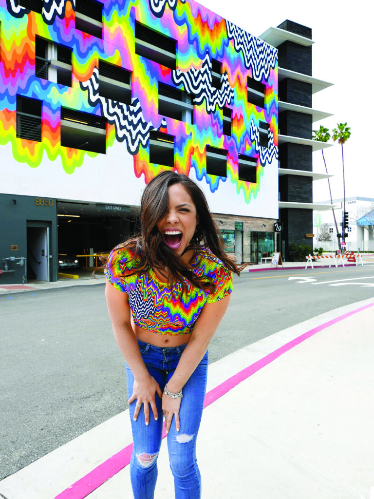 a colorful wavy rainbow stripe and black and white stripe crop top on a happy, excited woman matches a mural painted on a wall behind her