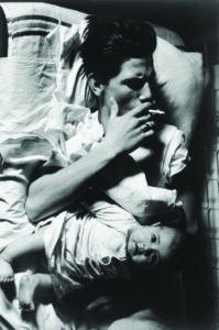 Black-and-white photograph of an adult reclining on a rumpled bed while holding a cigarette, with an infant lying across the adult's lap and looking toward the camera.