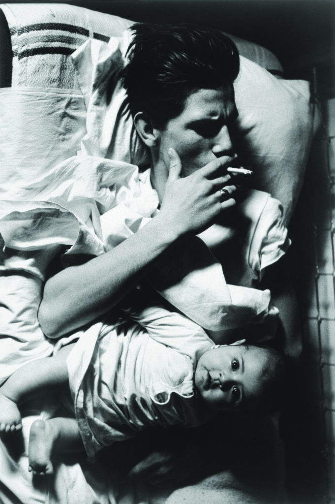 Black-and-white photograph of an adult reclining on a rumpled bed while holding a cigarette, with an infant lying across the adult's lap and looking toward the camera.