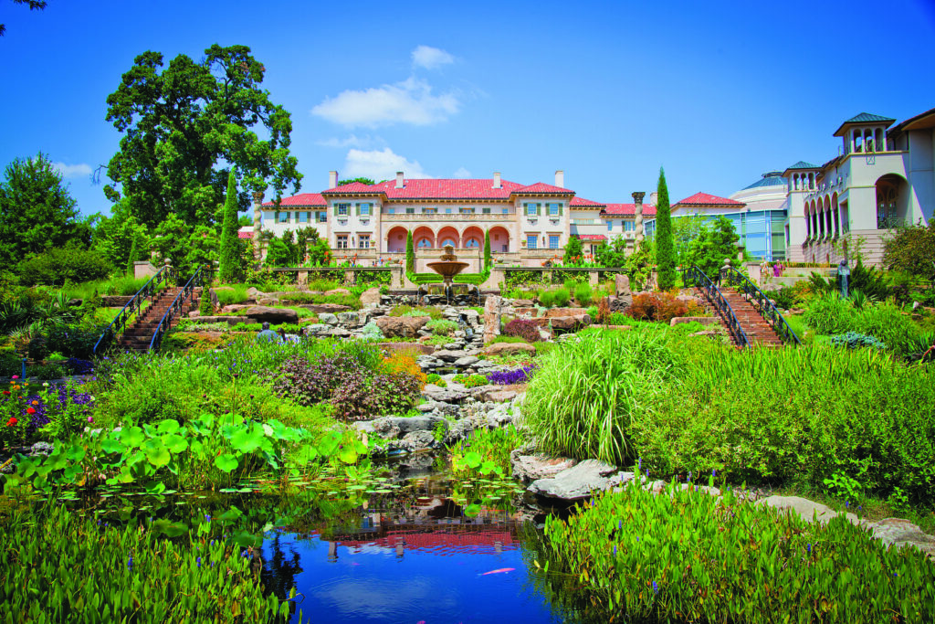 Large Mediterranean-style building with a red tile roof overlooking formal gardens, terraced landscaping, fountains, and a reflective pond under a bright blue sky.