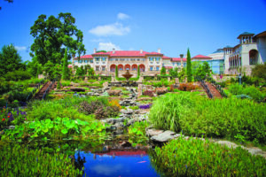 Large Mediterranean-style building with a red tile roof overlooking formal gardens, terraced landscaping, fountains, and a reflective pond under a bright blue sky.
