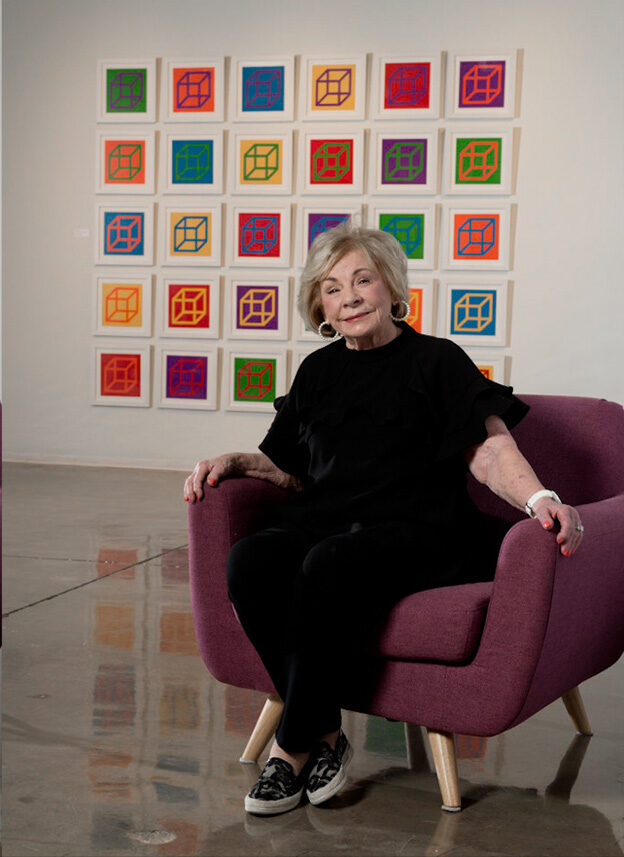 Portrait of Joan Maguire, a woman sits in an oversized chair in front of an art installation