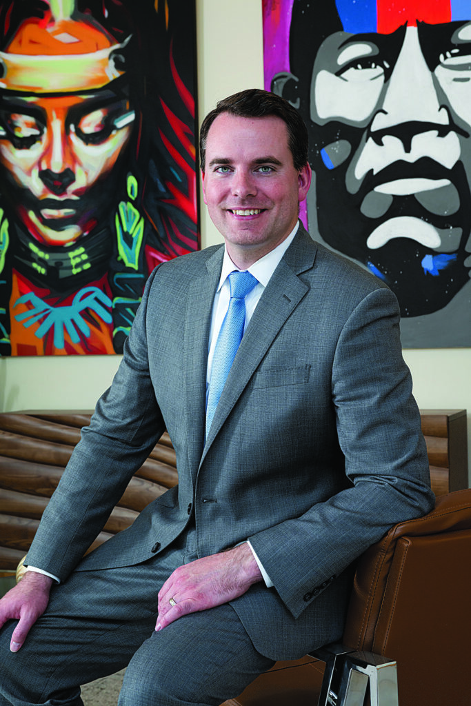 Portrait of Nathaniel Harding, a man in a suit who sits on a desk with artworks on a wall behind the desk