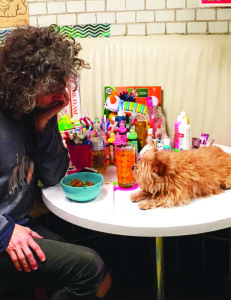 A man sits at a small table with his head in one hand as he looks a small dog laying on top of the table